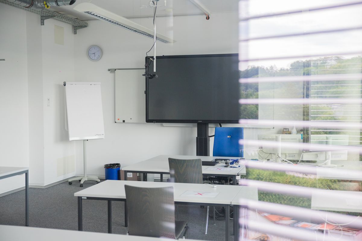 Blick durch ein Fenster in einen Unterrichtsraum der DAA Fachschule für Sozialpädagogik Aalen. Zu sehen sind einige Tische, eine digitale Tafel, sowie ein Flipchart. Im rechten Bereich reflektiert die Fensterscheibe die Umgebung der Schule.